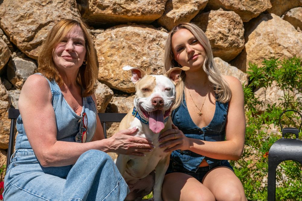 Two women sitting outdoors with a happy white pit bull dog, posing in front of a stone wall.