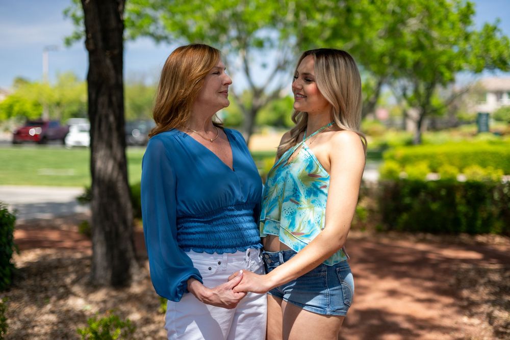 Two women holding hands and smiling at each other in a sunny park, wearing colorful summer outfits.