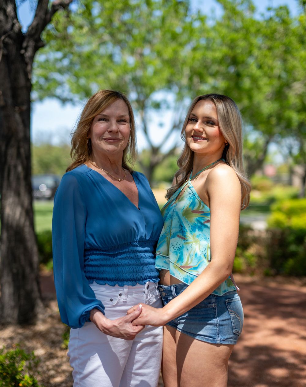 Two women, one in blue top and one in teal crop top, pose together in sunny park.