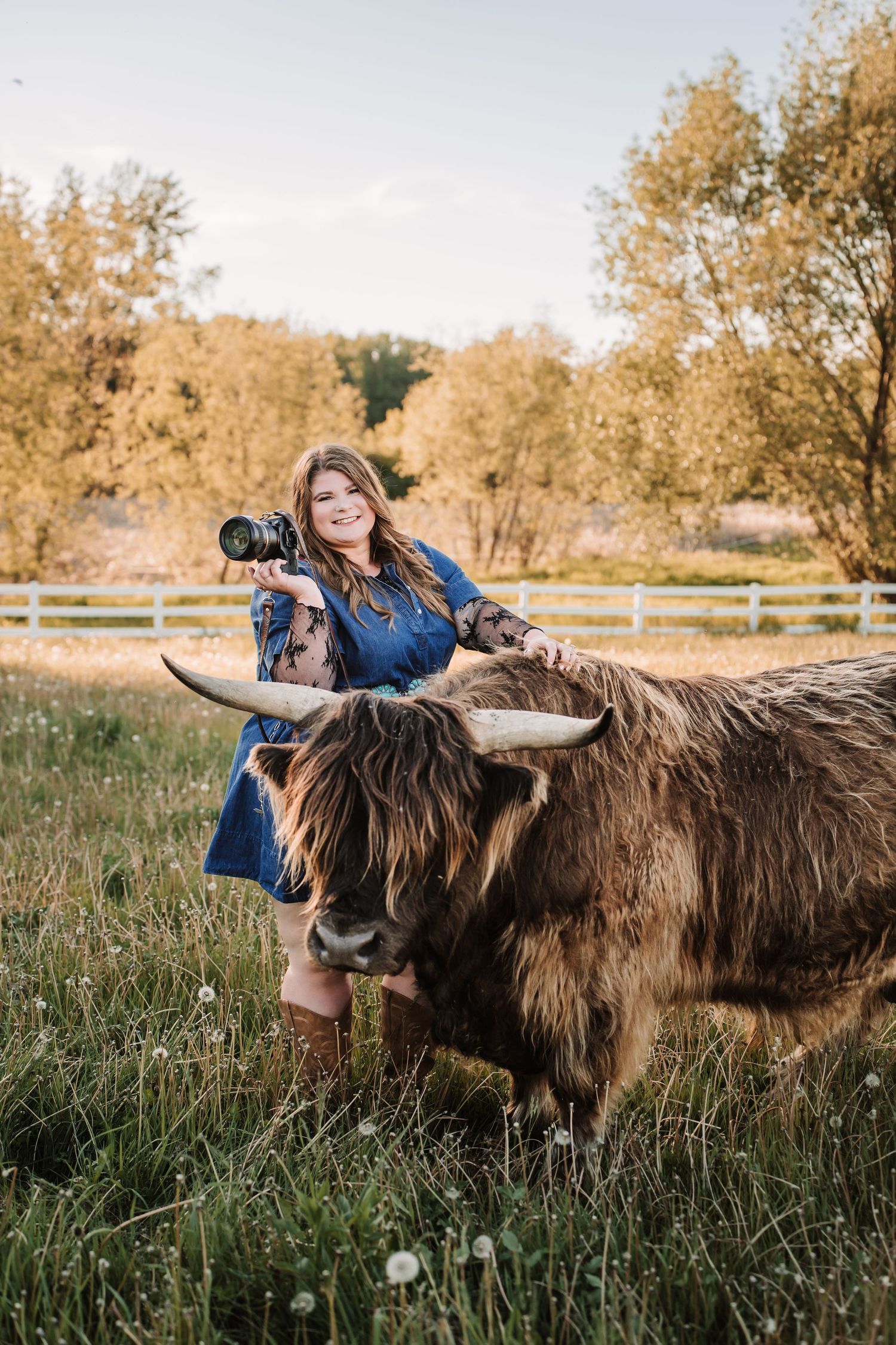 Smiling woman in blue dress holding camera poses with a large Highland cow in a green pasture with white fence.