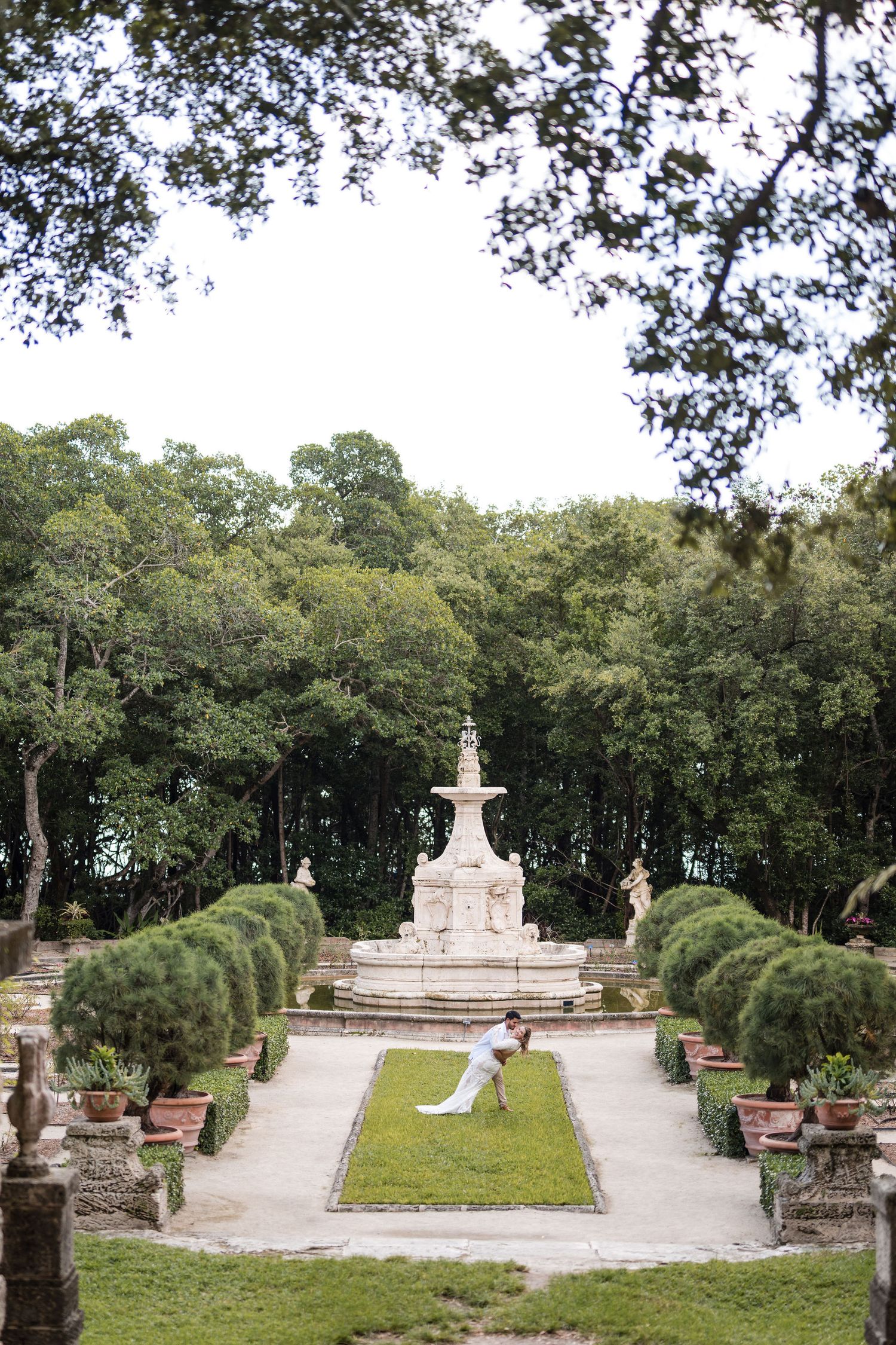 Bride and groom walk hand in hand through Vizcaya Gardens, framed by stone pathways, manicured hedges, and a historic fountain.