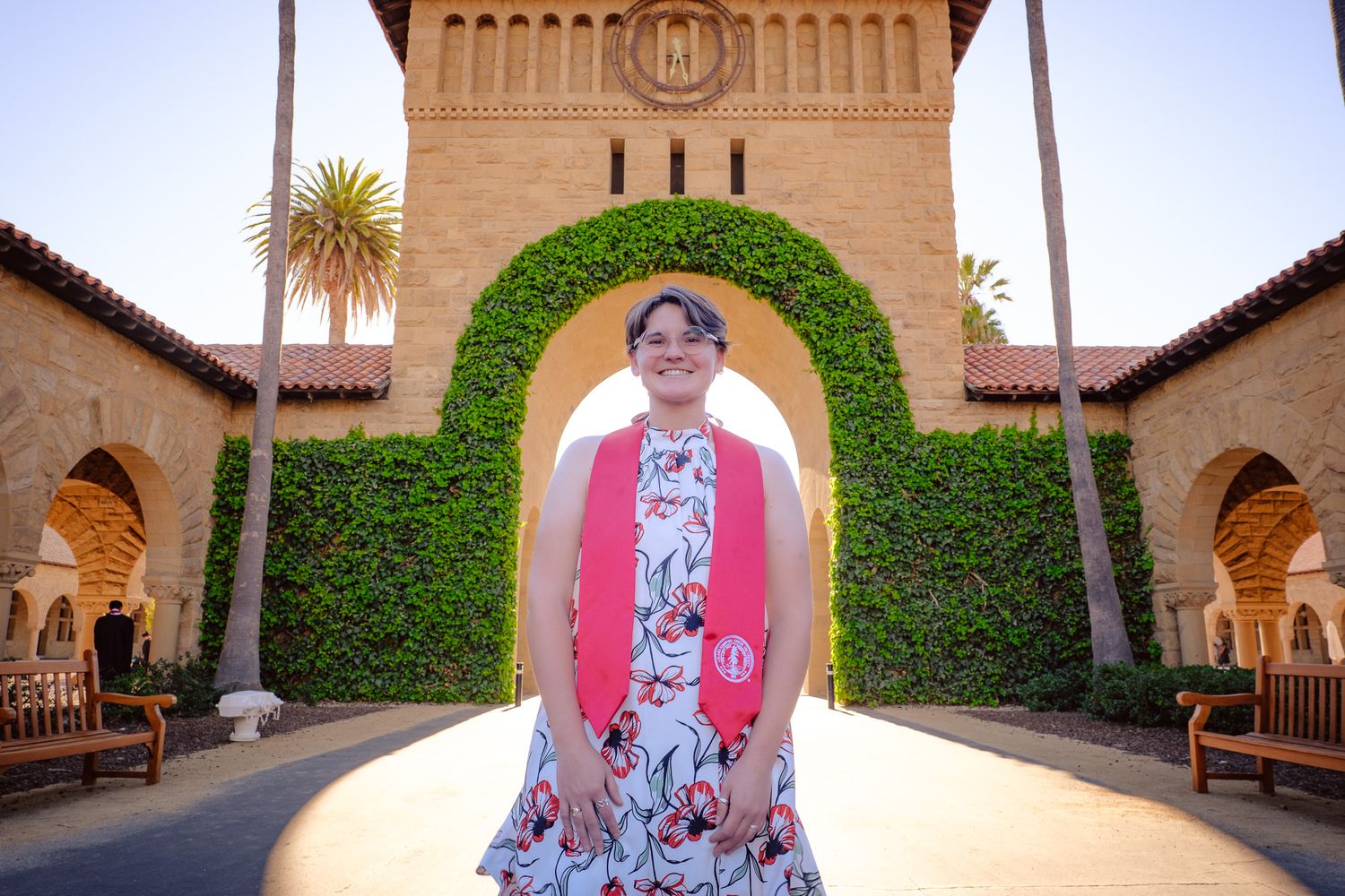 Graduate in floral dress with pink sash poses under ivy arch at Stanford University campus on sunny day.