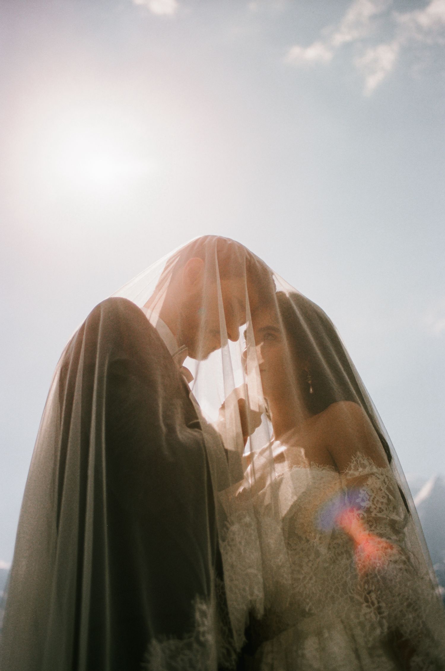 A bride and groom share an intimate moment under a flowing veil, backlit by soft sunlight against a blue sky.