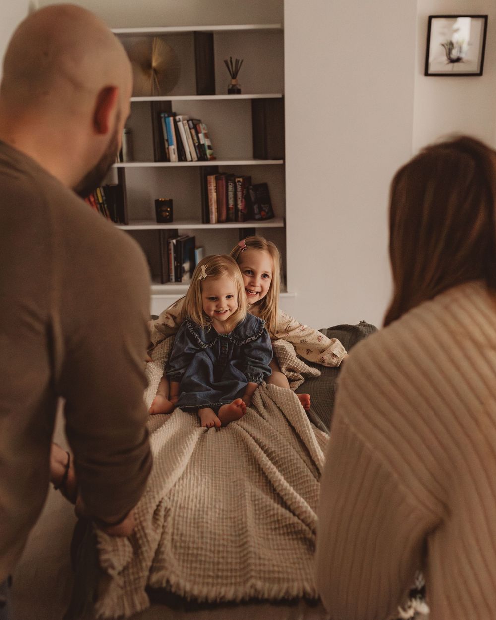 Two young sisters smiling under a blanket on a bed as parents look on lovingly.