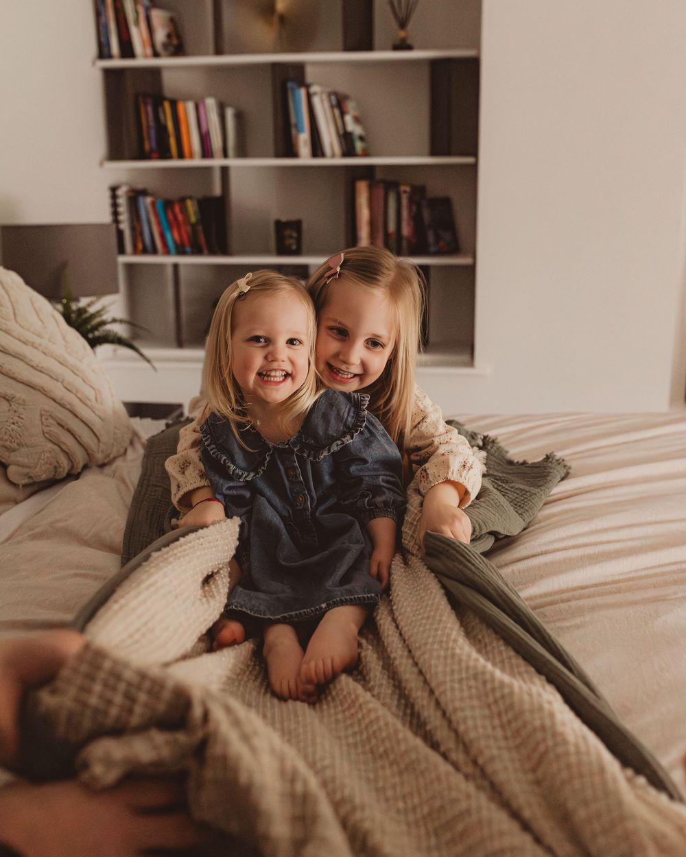 Two smiling blonde sisters sitting together on a bed with neutral bedding in a cozy bedroom.