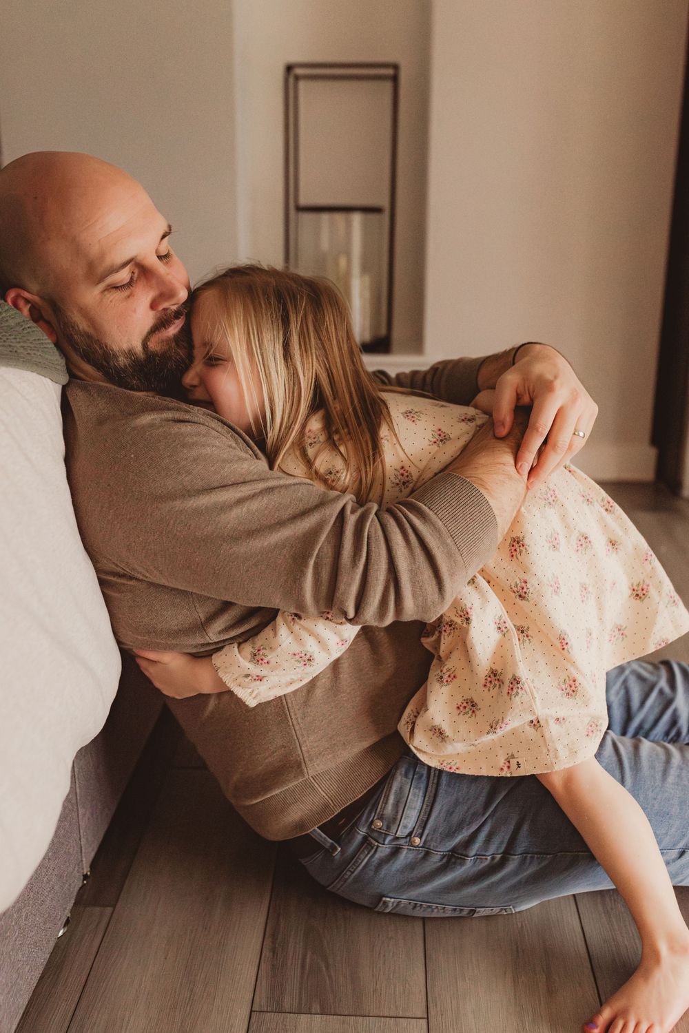 Bald father tenderly hugging his young blonde daughter while sitting on bedroom floor.