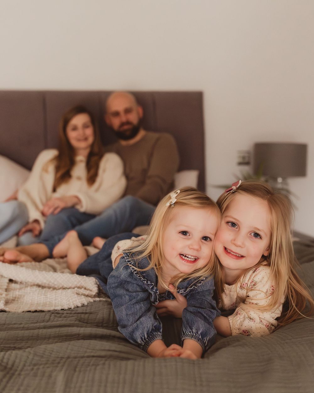 Family portrait with two blonde girls lying on bed smiling while parents sit behind them.