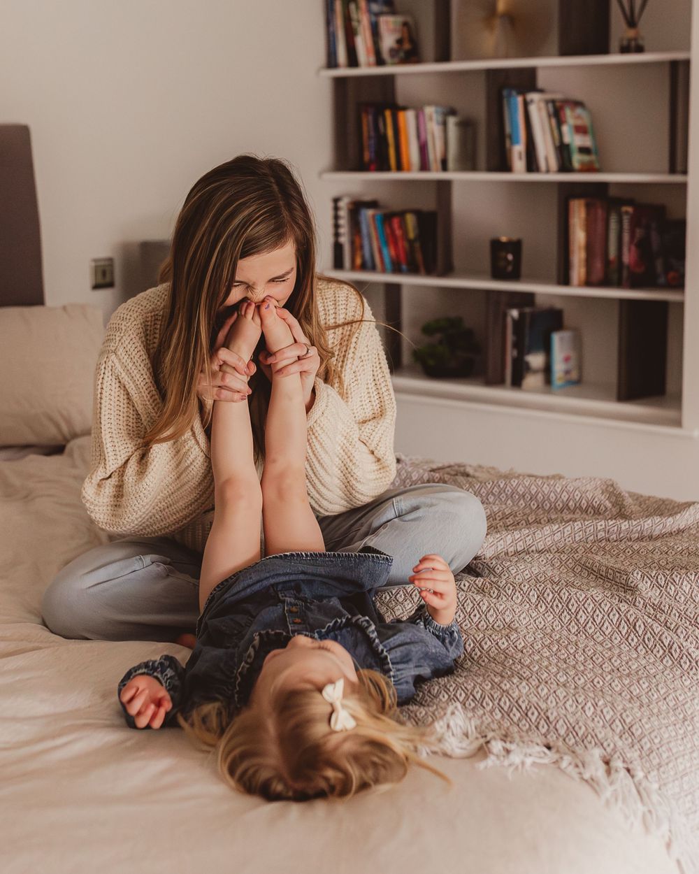 Mother playfully kissing toddler feet while child lies on neutral-toned bed laughing.