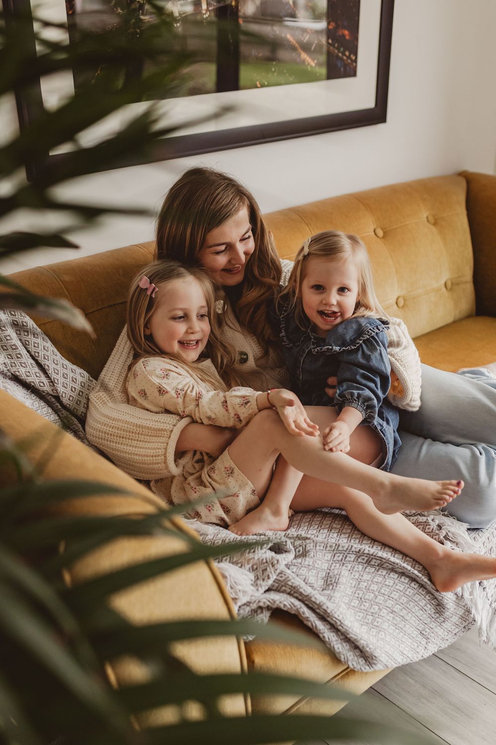 Happy mother laughing with two young daughters snuggled on a cozy yellow couch indoors.