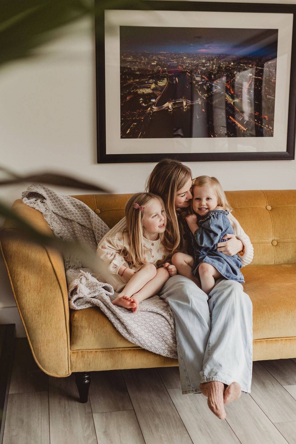 Mother sitting on yellow sofa cuddling two young daughters with a city night photo on wall.
