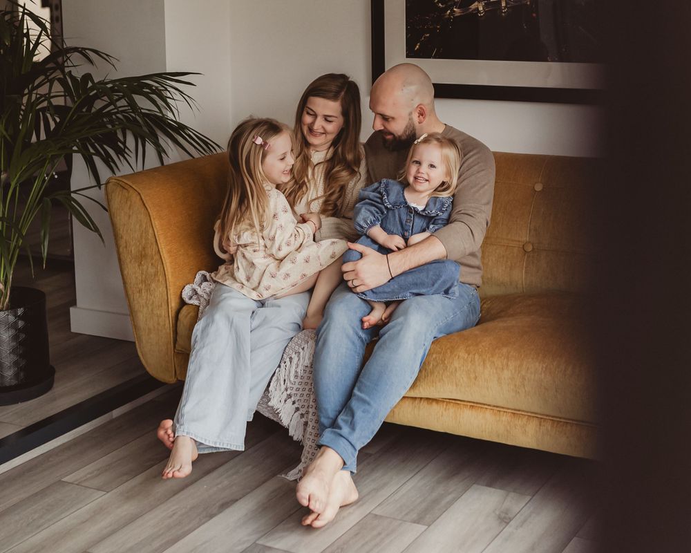 Smiling family with two young daughters sitting barefoot on a golden velvet sofa indoors.