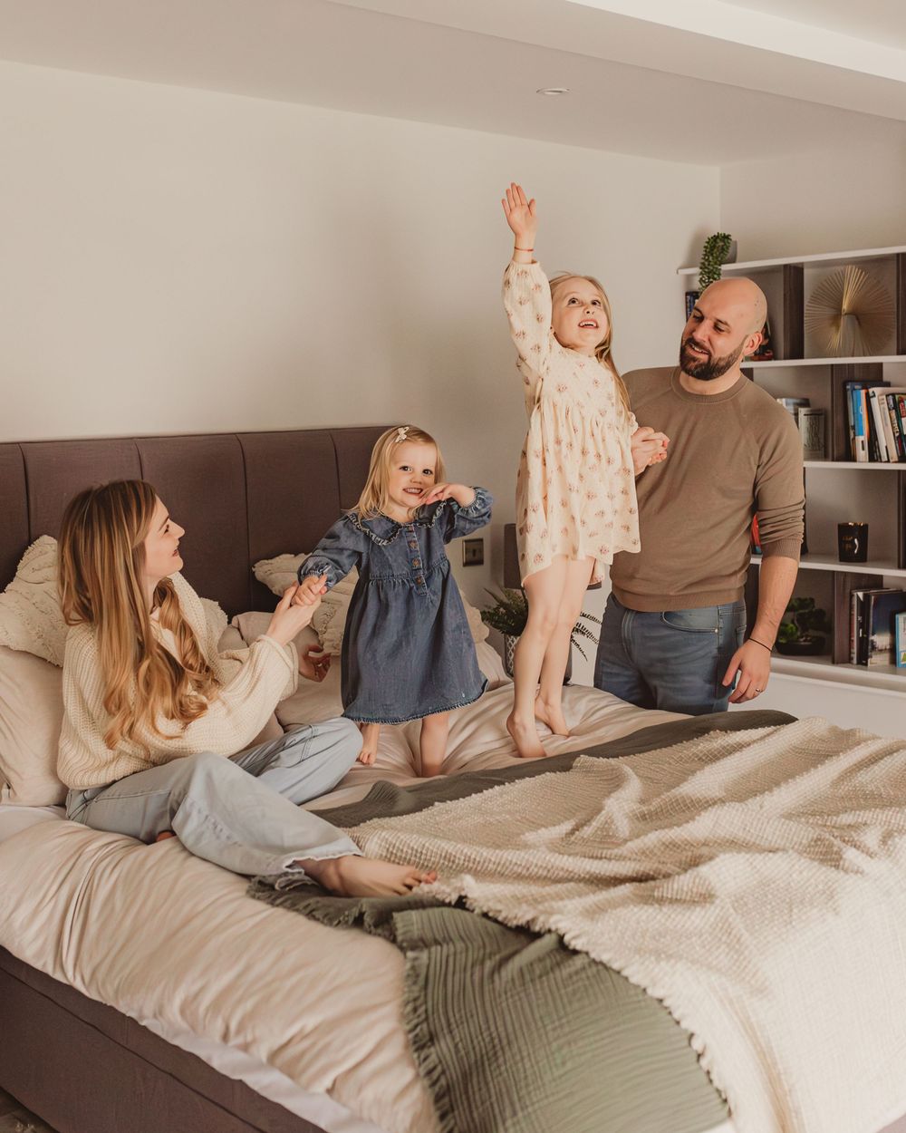 Joyful family watching their young daughter jump on a bed in a bright modern bedroom.