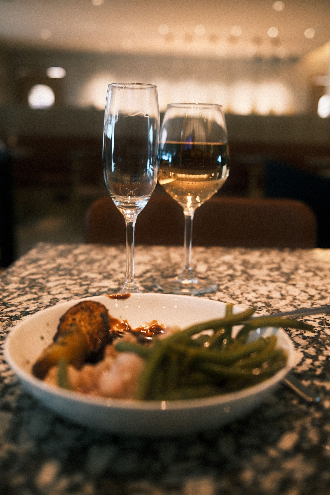 Elegant restaurant table with two wine glasses and a white plate of green beans, shrimp, and baked dish on granite surface.