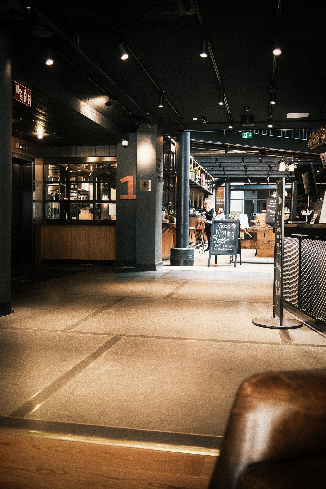 Modern industrial bar interior with dark ceiling, warm lighting, wooden counters, and a chalkboard sign on polished concrete floors.