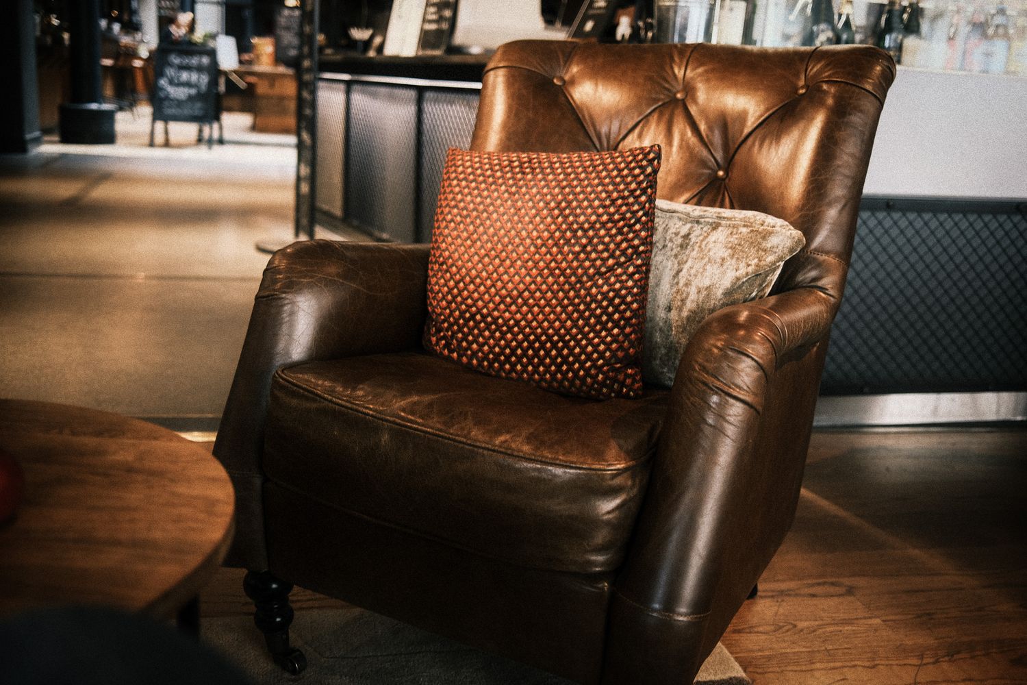 Vintage brown leather tufted armchair with decorative cushions on hardwood floor in cozy interior setting.
