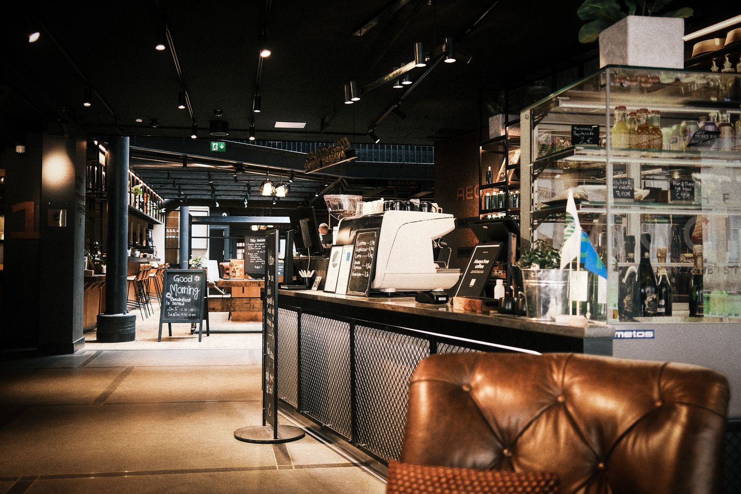 Modern industrial-style coffee bar with leather seating, espresso machine, and dark interior lighting.