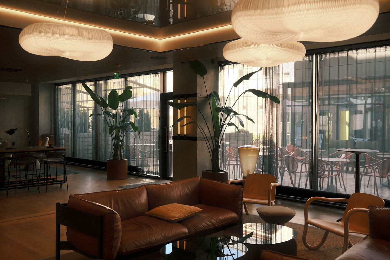 Modern hotel lobby with leather sofas, pendant lights, glass walls, and tropical plants in warm ambient lighting.