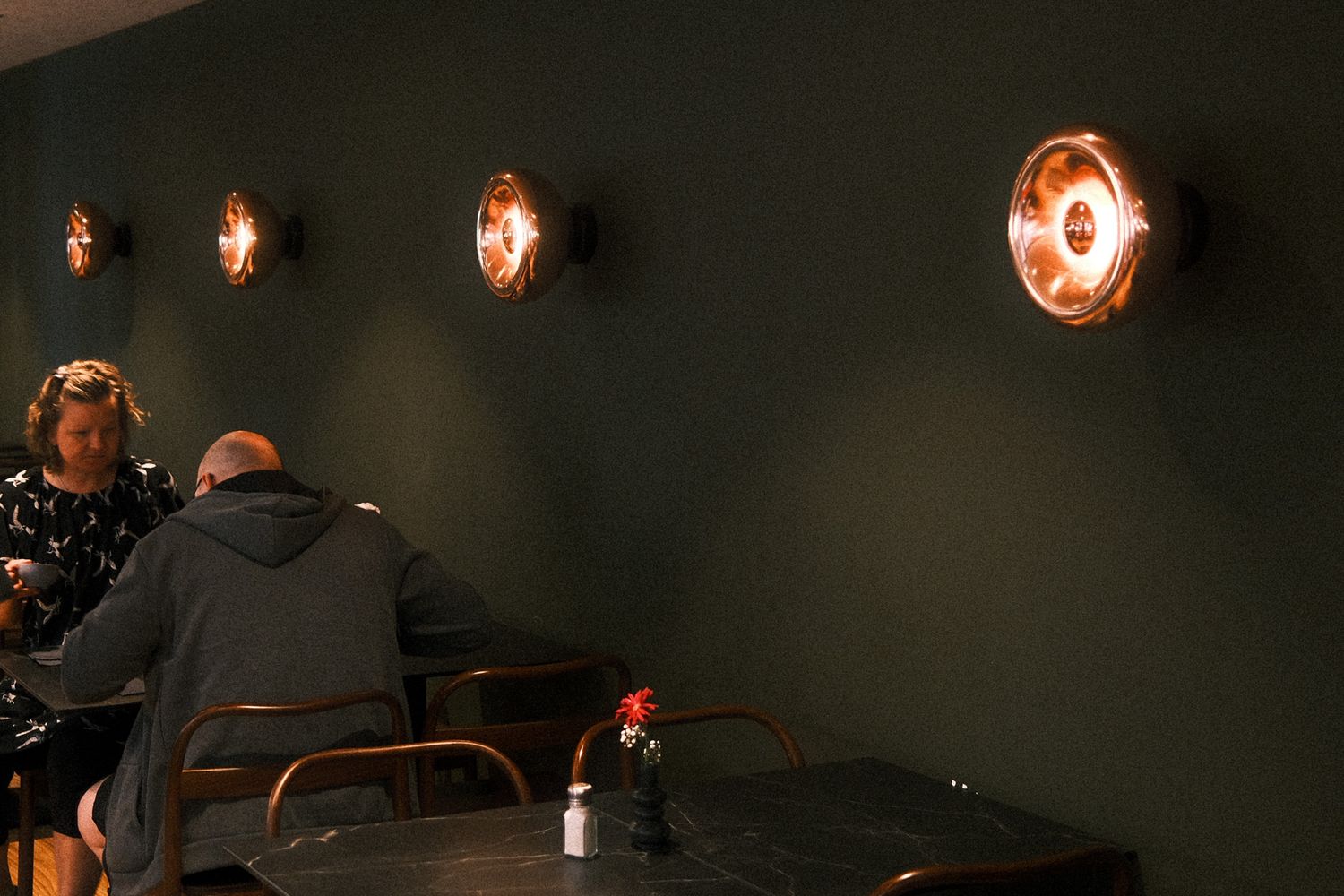 Two diners sit beneath warm copper wall sconces against a dark green wall in a cozy restaurant setting.