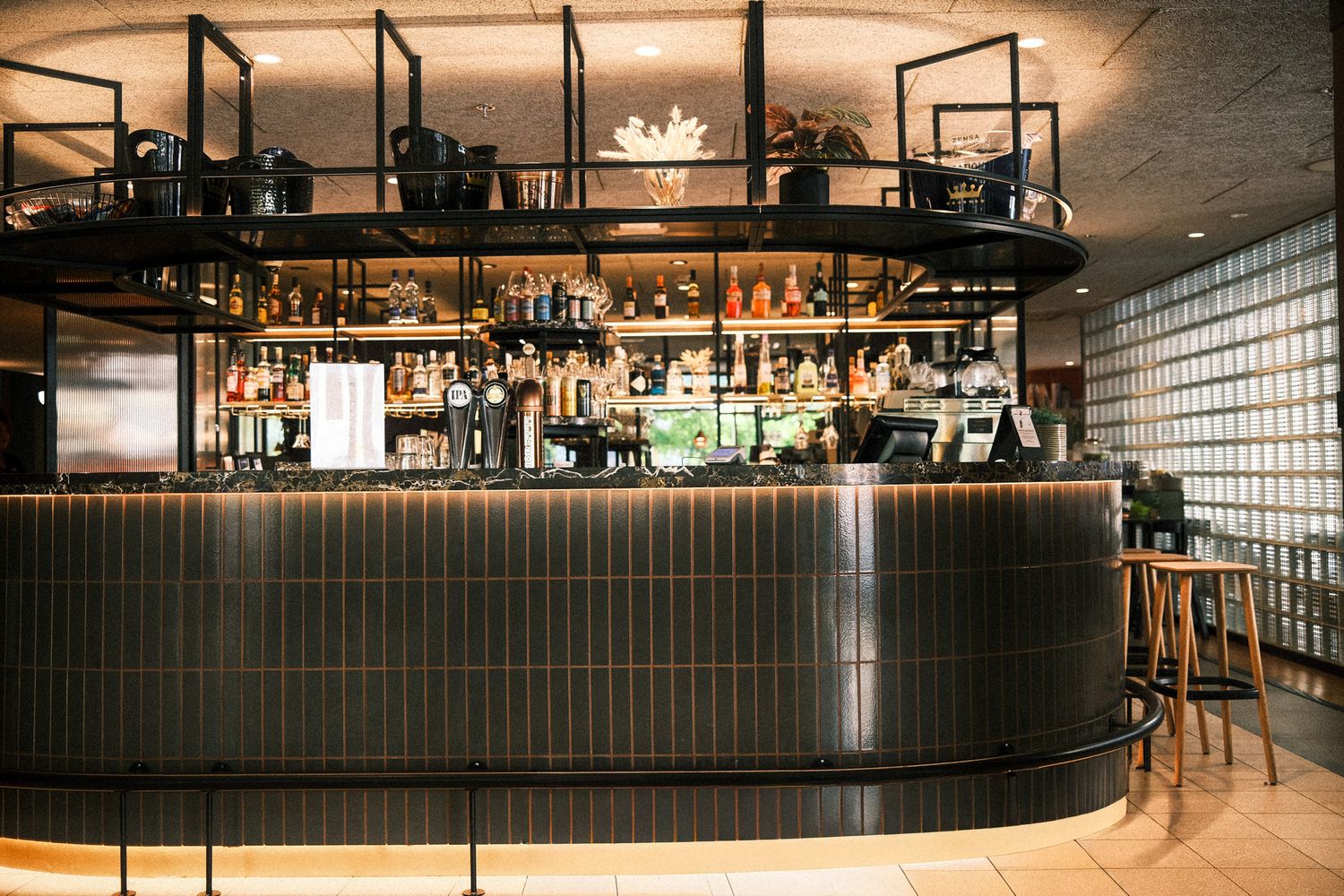 Elegant curved bar with dark tiled facade, backlit shelves of spirits, and wooden stools in a modern restaurant.
