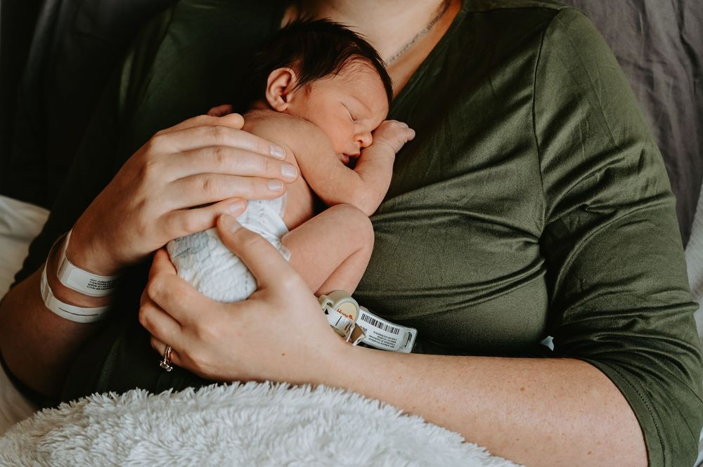 Mother in green shirt cradling sleeping newborn baby against her chest in a tender postpartum moment.