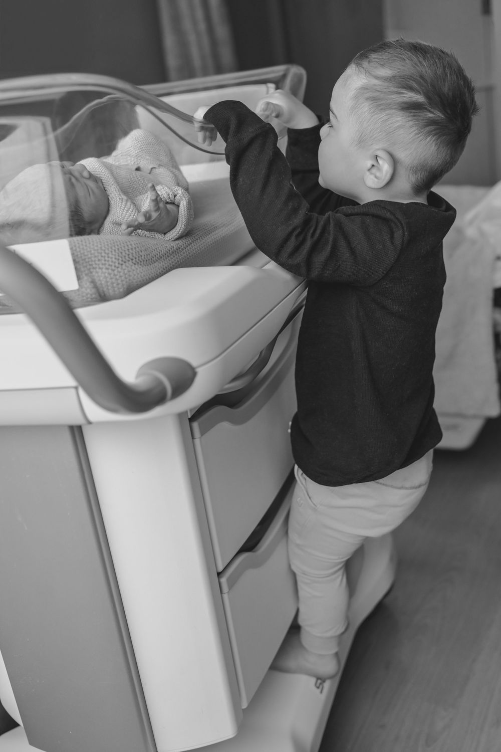 Black and white photo of a young boy standing on tiptoes to peek at a newborn baby in a hospital bassinet.