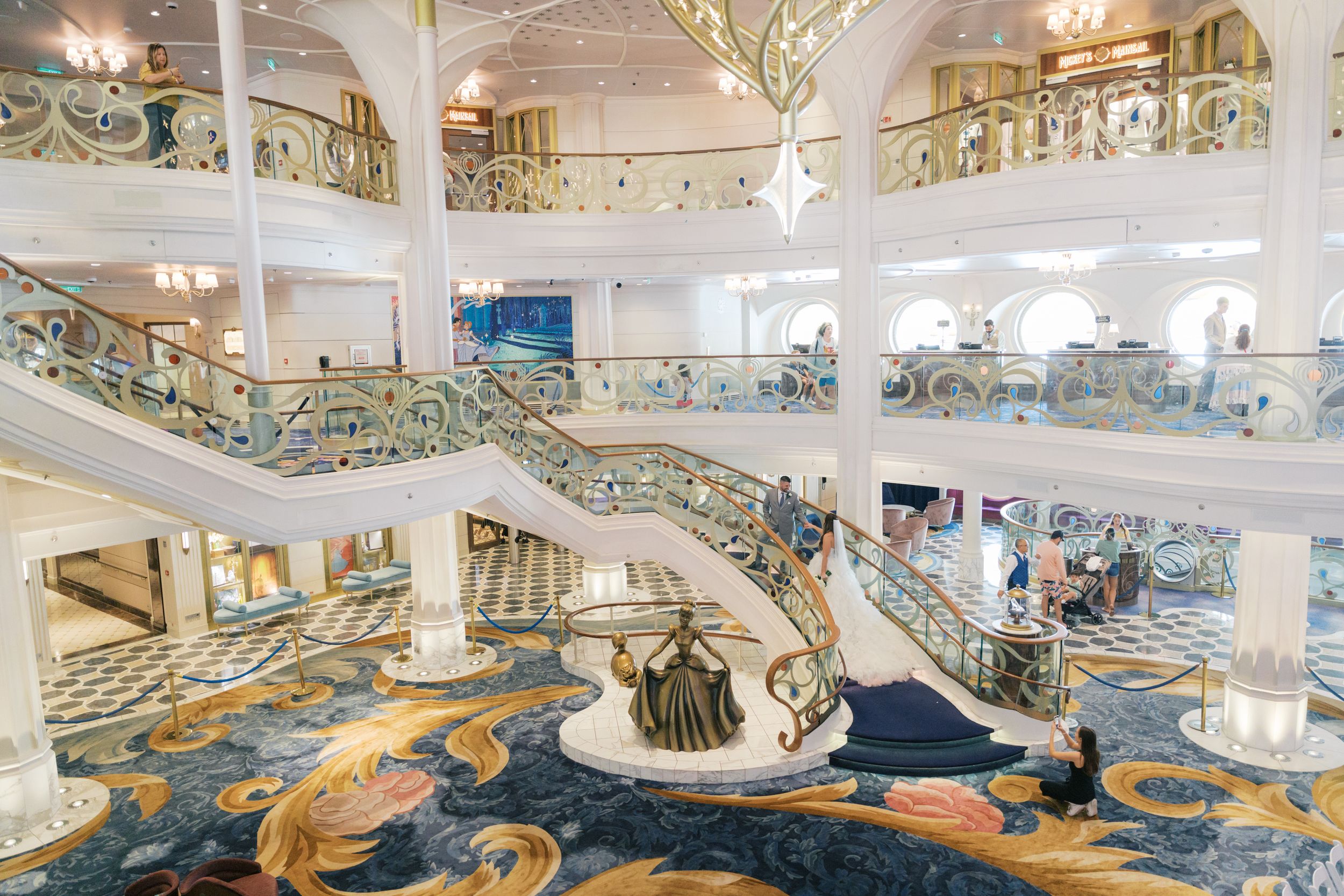 Elegant cruise ship atrium with grand sweeping staircases, ornate gold railings, and colorful mosaic carpet flooring.