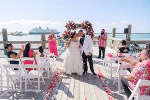 Bride and groom kiss on a waterfront deck wedding ceremony with floral arch and rose petals, cruise ship in background.