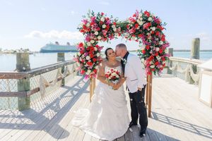 Bride and groom kissing under a heart-shaped floral arch on a waterfront dock with a cruise ship in the background.