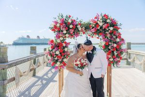Bride and groom kissing under a vibrant heart-shaped floral arch on a dock with a Disney cruise ship behind them.