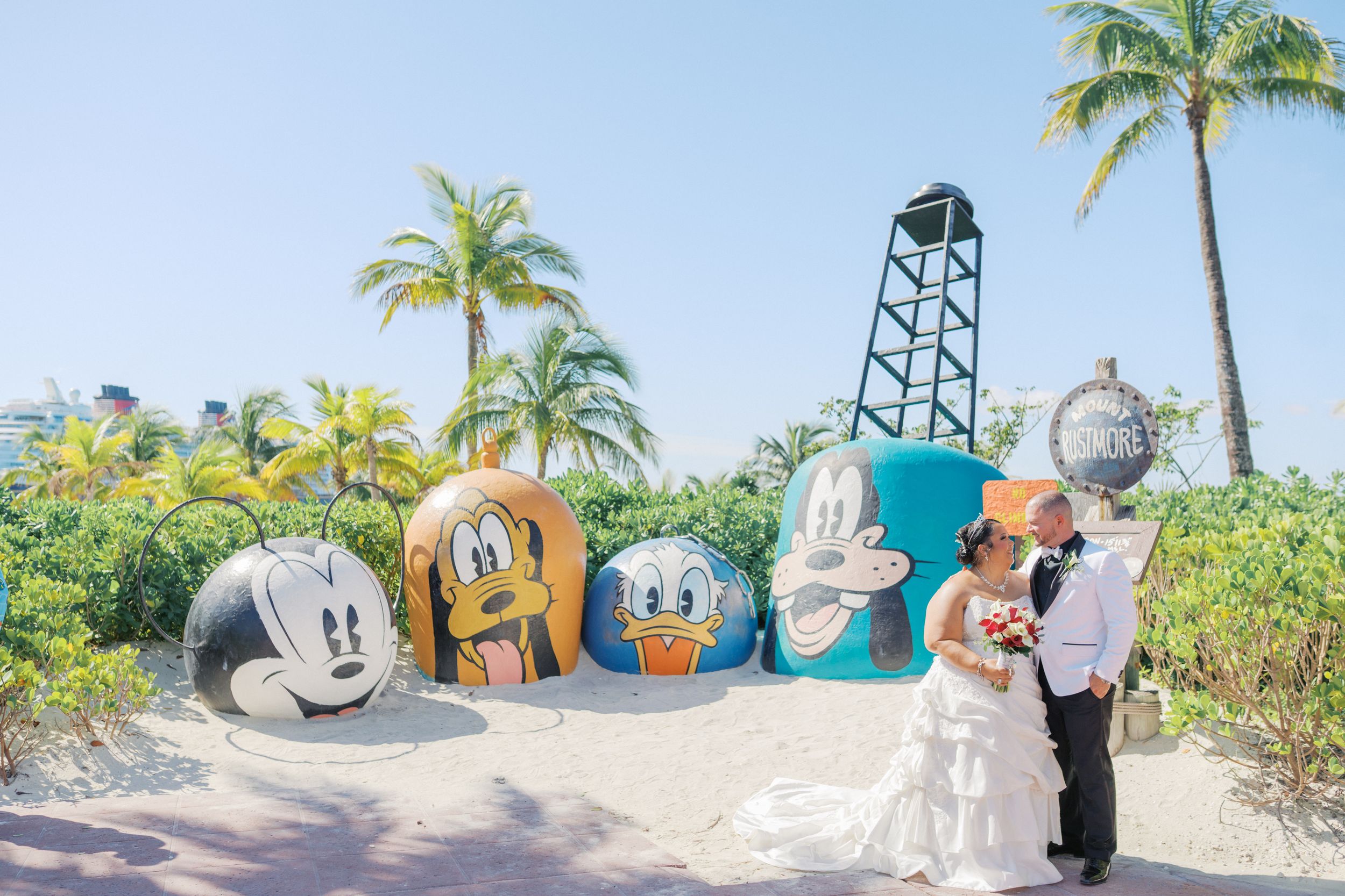 Newlyweds kissing in front of colorful Disney character rocks on a tropical beach with palm trees.