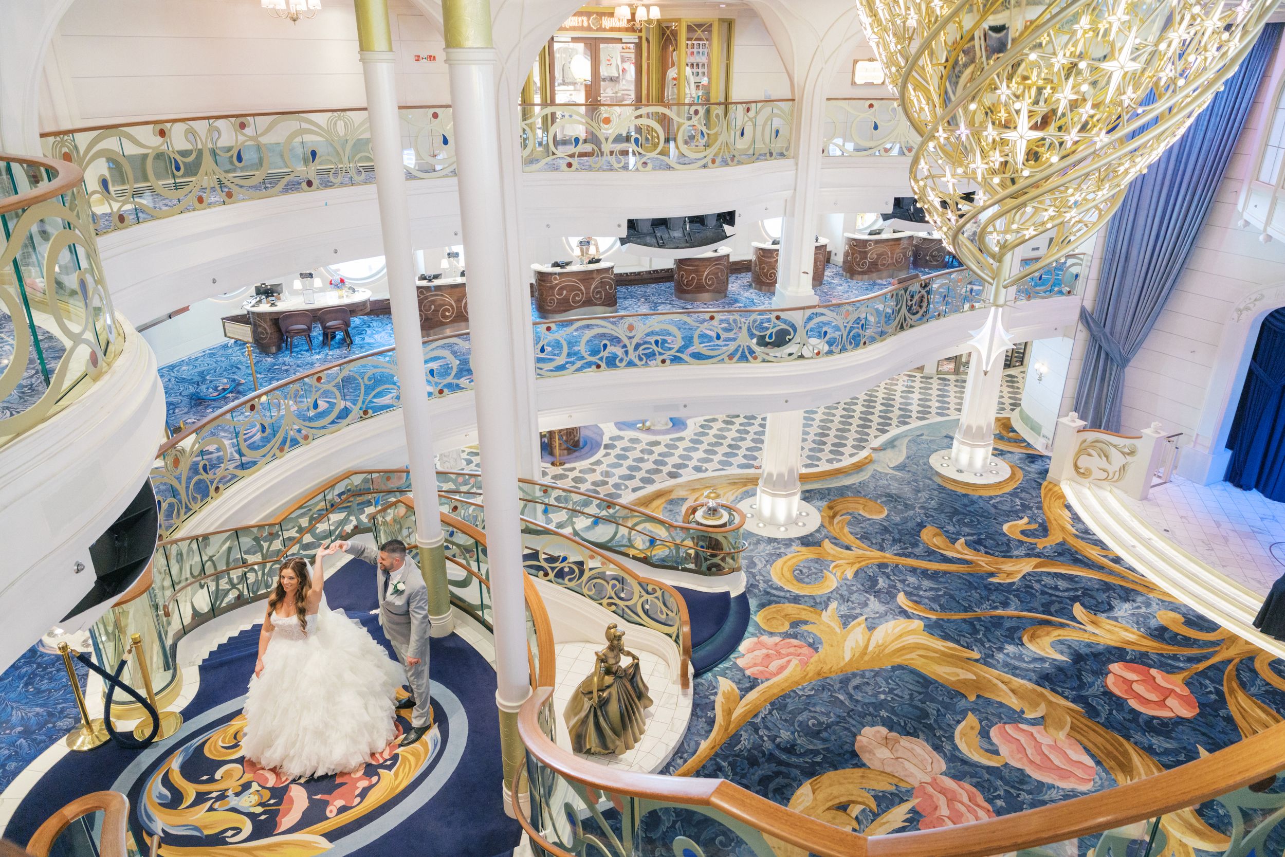 Bride in white gown poses on ornate spiral staircase in luxurious blue and gold ballroom.