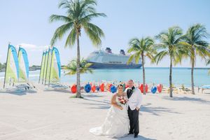 Bride and groom pose on a tropical beach with palm trees and a Disney cruise ship in the background.