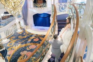 Groom leads bride down elegant spiral staircase in opulent blue and gold decorated venue.