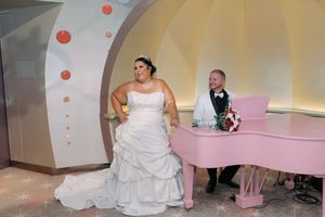 Bride in white gown stands beside groom seated at pink baby grand piano during elegant wedding reception.