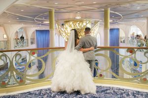 Bride in white ballgown and veil stands with groom overlooking elegant cruise ship atrium balcony.