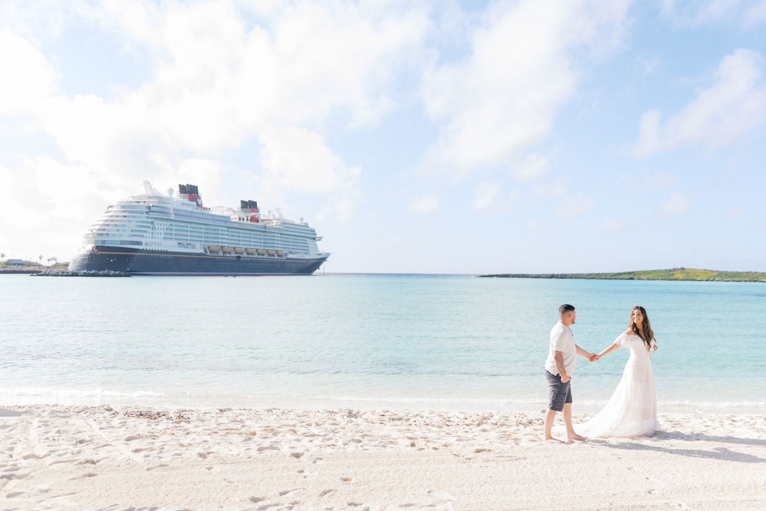 Couple holding hands on a white sand beach with a Disney cruise ship in the turquoise waters behind them.