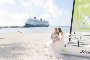 Couple in wedding attire sit by a sailboat on a tropical beach with a cruise ship in the background.