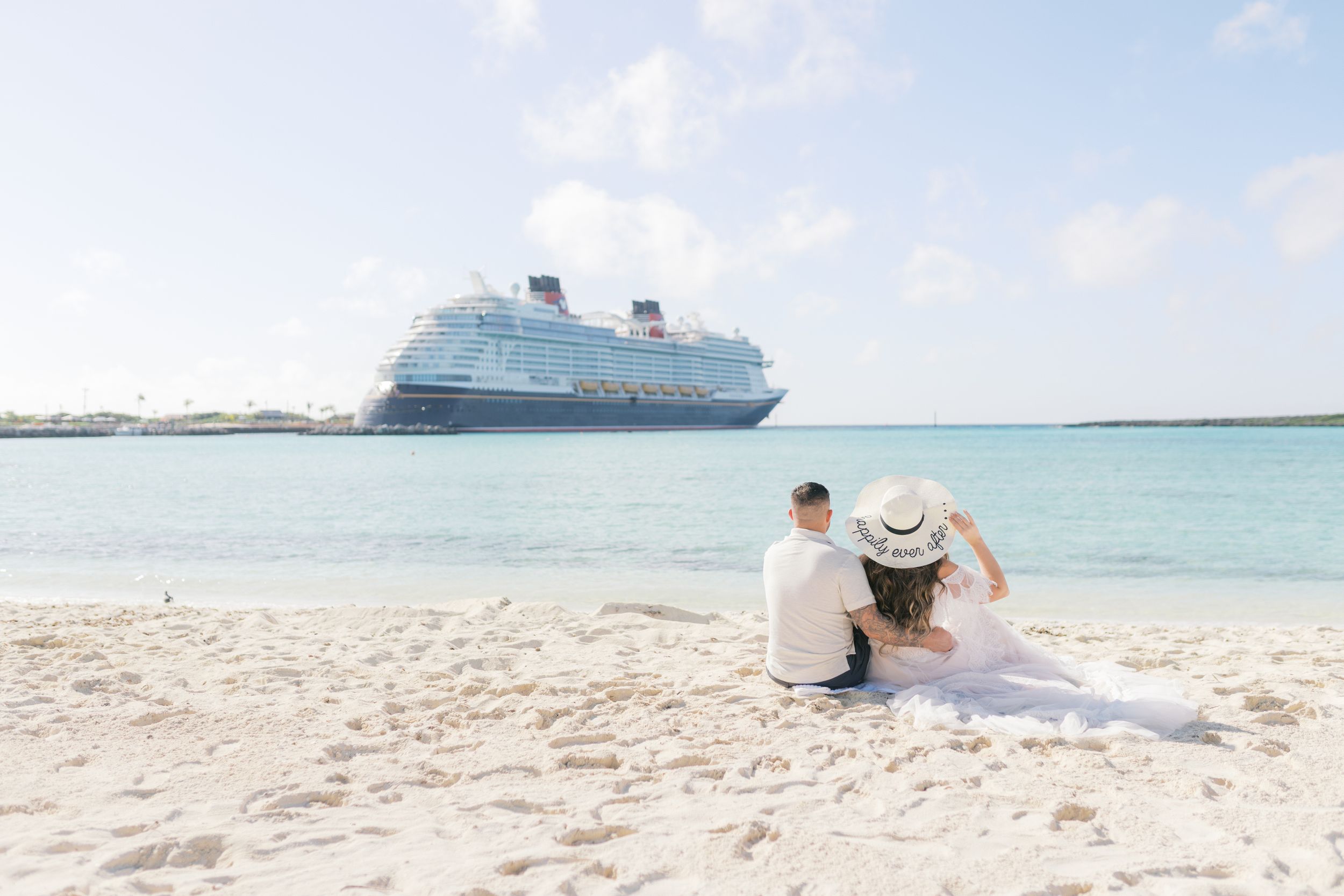 Bride and groom sit on white sandy beach gazing at Disney cruise ship on horizon.