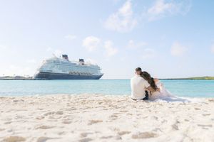 Couple in wedding attire sitting on white sandy beach with a Disney cruise ship in the turquoise ocean backdrop.