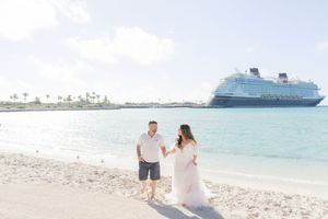 Couple walking on a white sand beach with a Disney cruise ship in the background on a sunny day.