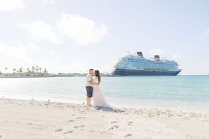 Newlyweds share a romantic kiss on a pristine beach with a large cruise ship anchored in the distance.
