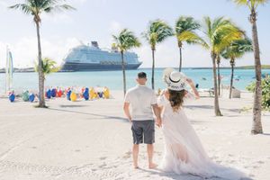 Couple holding hands on tropical beach with cruise ship and palm trees in background.