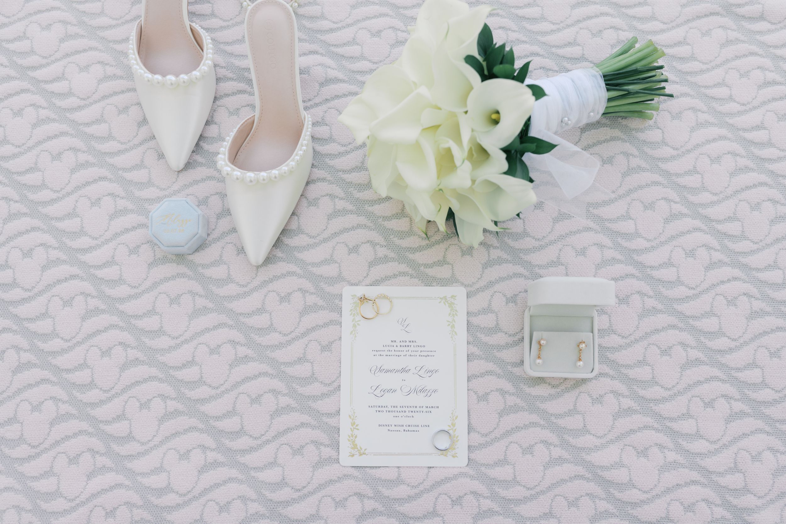 White wedding heels, calla lily bouquet, elegant invitation, ring box, and pearl earrings arranged on a lace textured surface.