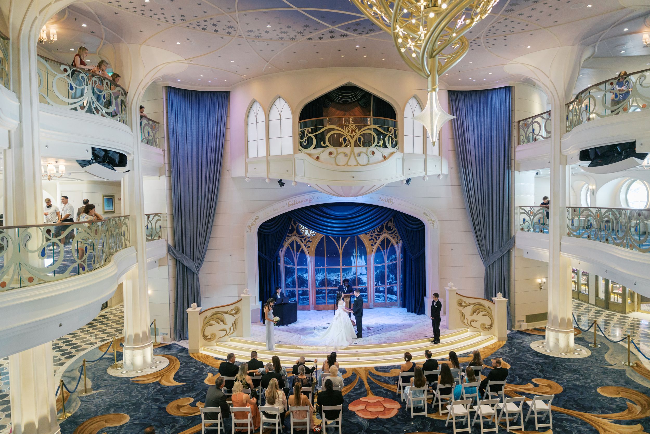 Elegant Disney cruise ship atrium wedding ceremony with blue curtains, grand staircase, and guests seated on ornate patterned carpet.