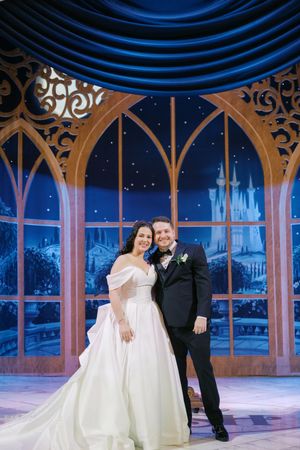 Bride and groom pose together in elegant wedding attire before a fairytale castle backdrop with blue drapes.