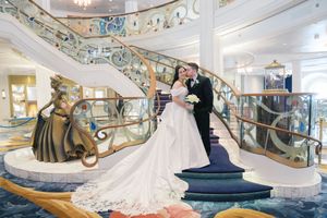 Bride and groom share a kiss on a grand staircase aboard a Disney cruise ship, with an elegant golden statue nearby.