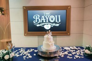 Three-tier wedding cake displayed at The Bayou venue with floral decorations and rose petals on a blue tablecloth.