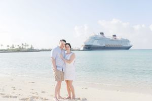 Couple smiling on a white sand beach with a Disney cruise ship docked in the turquoise water behind them.