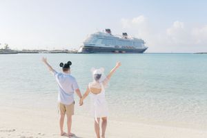 Couple holding hands on a tropical beach waving at a Disney cruise ship in the turquoise waters.