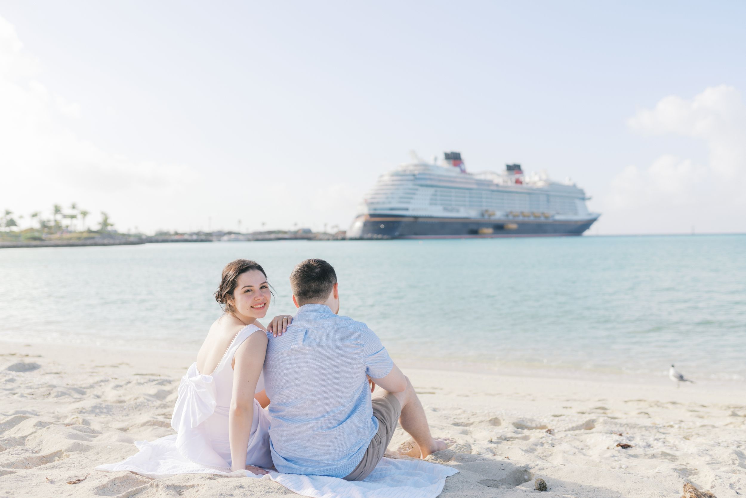 Couple sitting on a sandy beach in front of a Disney cruise ship, woman in white dress looking back over her shoulder.